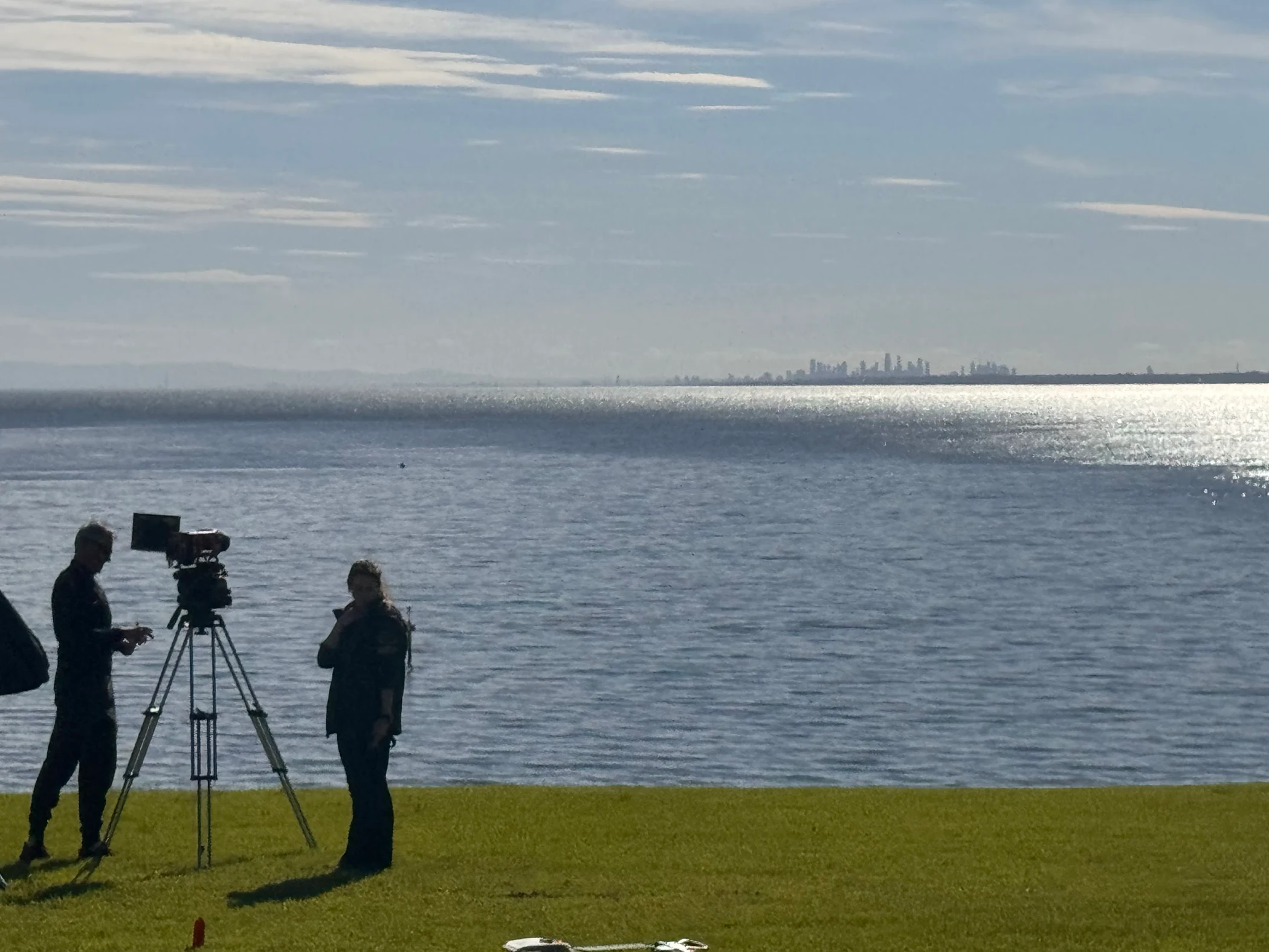 A crew working with a tripod-mounted cinema camera beside a waterfront, with a distant skyline.